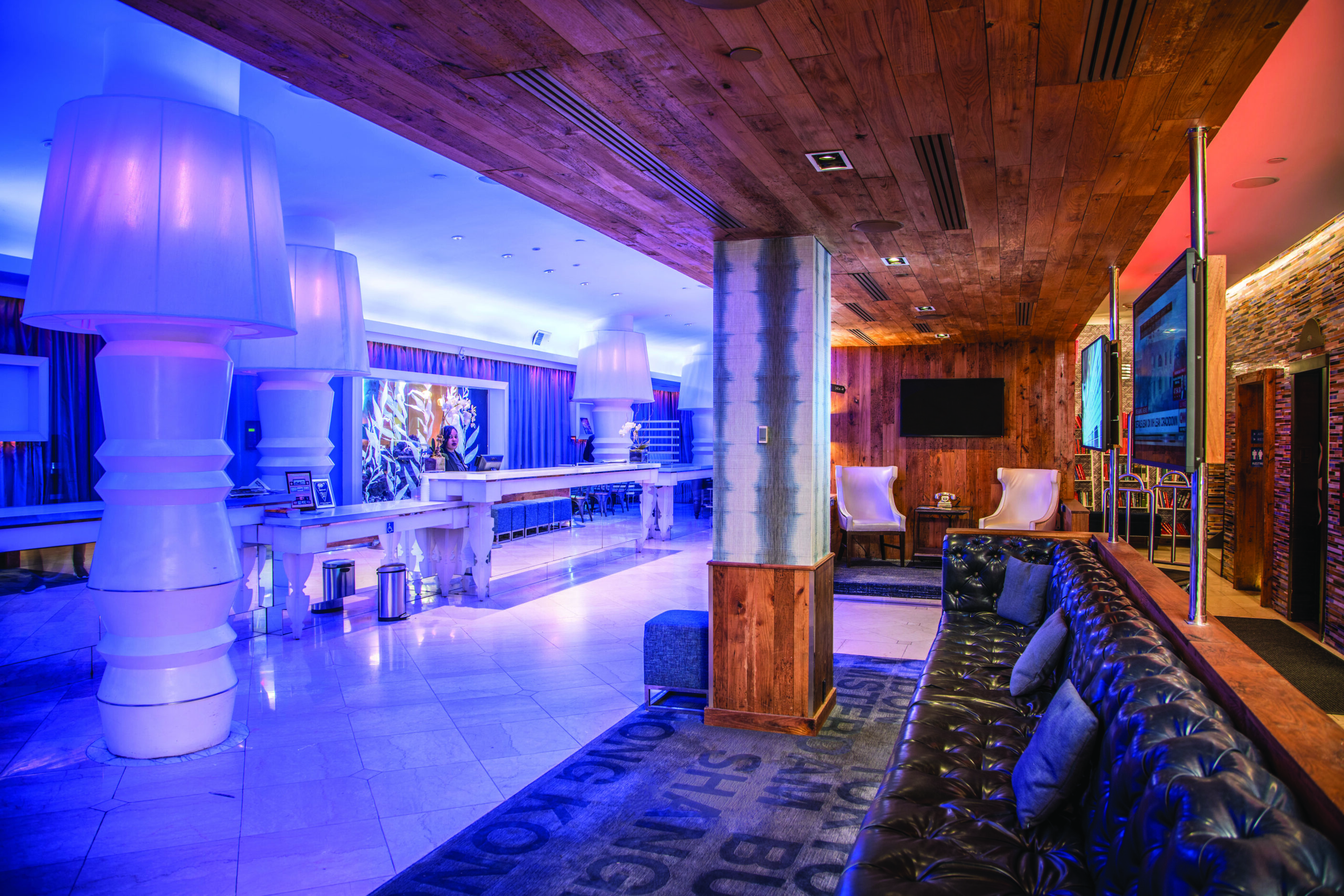Modern hotel lobby at Gallivant Times Square with blue and purple lighting, tall lamps, a sleek white bar, and black leather seating—an ideal Braintrust meeting spot.