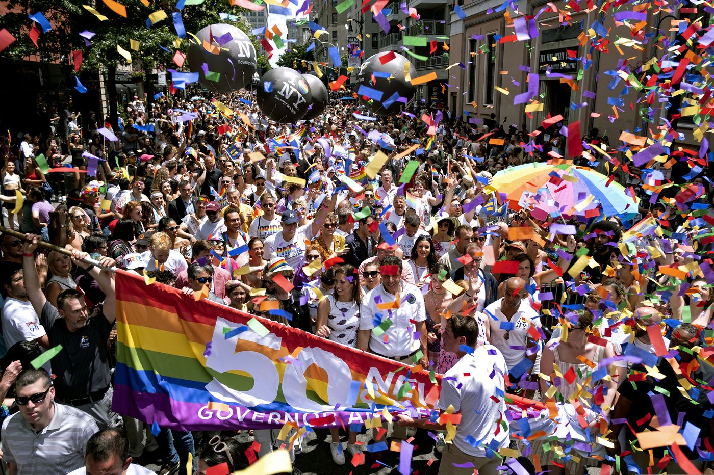 Crowd celebrating at a Pride parade with rainbow flags and confetti, holding a 50 NYC anniversary banner.
