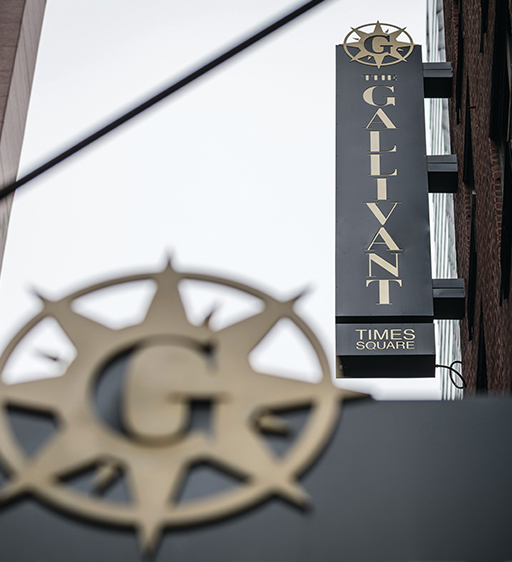 Vertical Braintrust Gallivant Times Square hotel sign with a compass logo, viewed from a low angle against the sky.