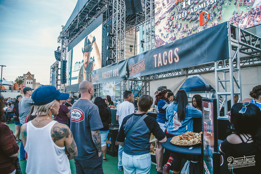 People line up at an outdoor taco stand at a lively Braintrust event at DLVEC, with a large stage and screen in the background.