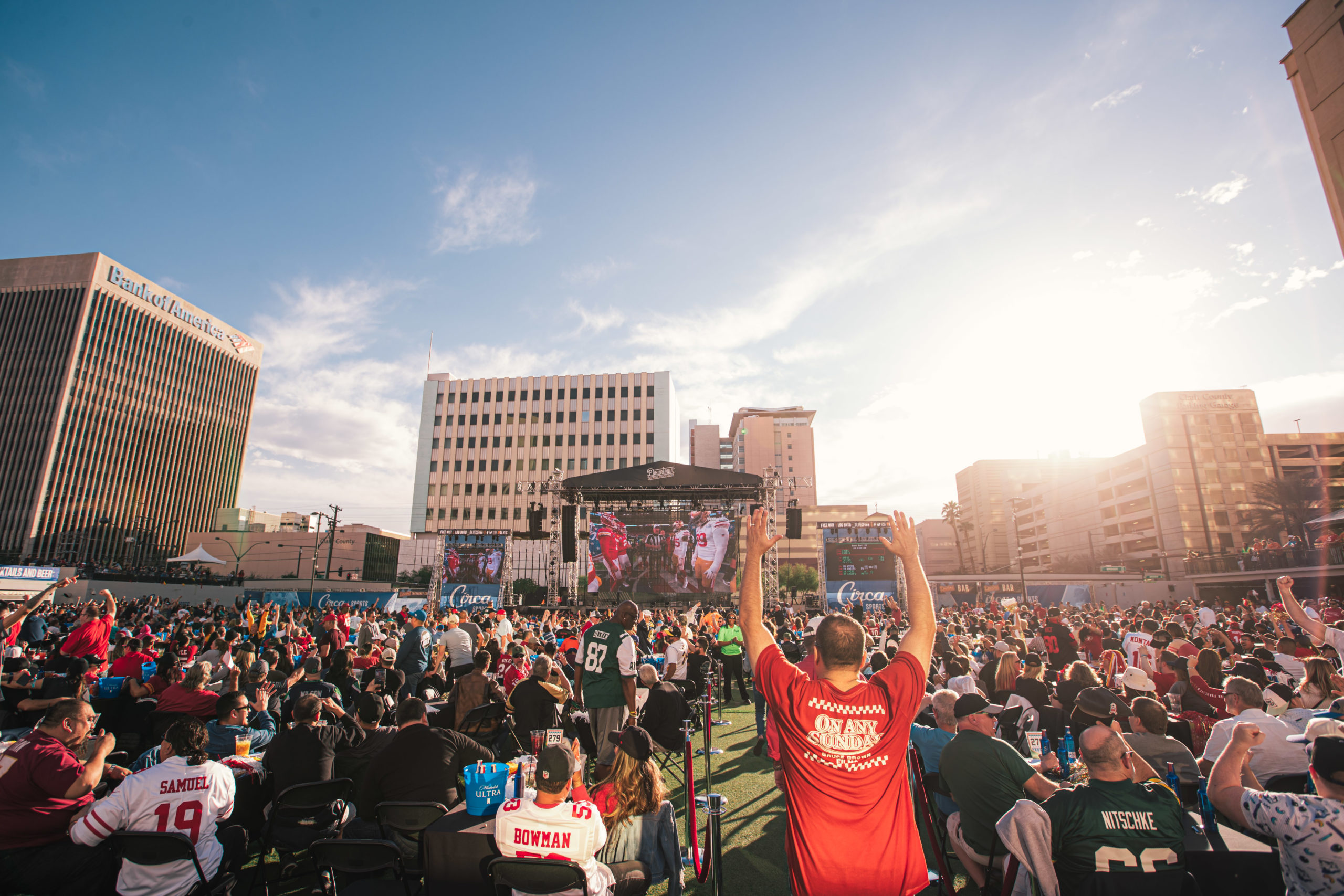 A large crowd gathers at DLVEC, watching a football game on a big outdoor screen in the city, sunlight streaming past tall buildings—a true Braintrust of fans united by their passion.
