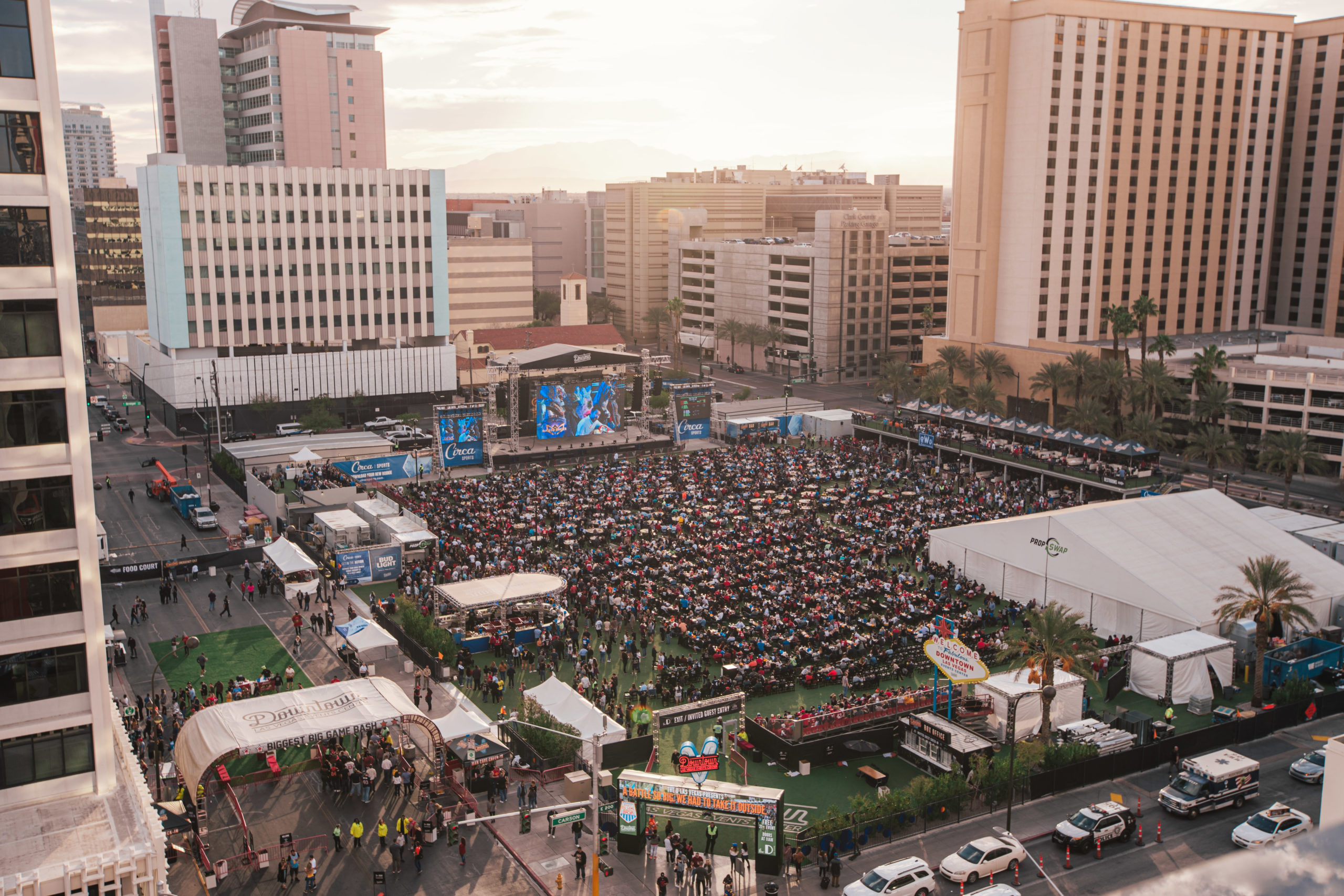 A large outdoor event at DLVEC, organized by Braintrust, with crowds seated in front of big screens in a city surrounded by tall buildings.