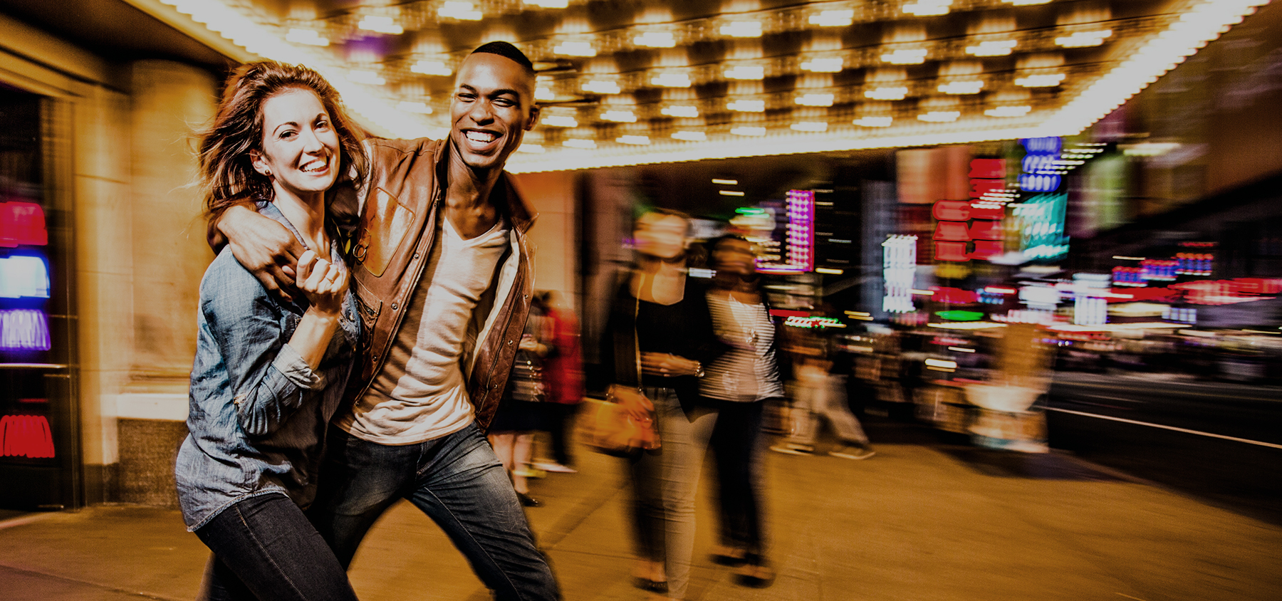 A smiling couple, part of the Braintrust, walks arm-in-arm under bright marquee lights, gallivanting through lively Times Square on a vibrant city night.
