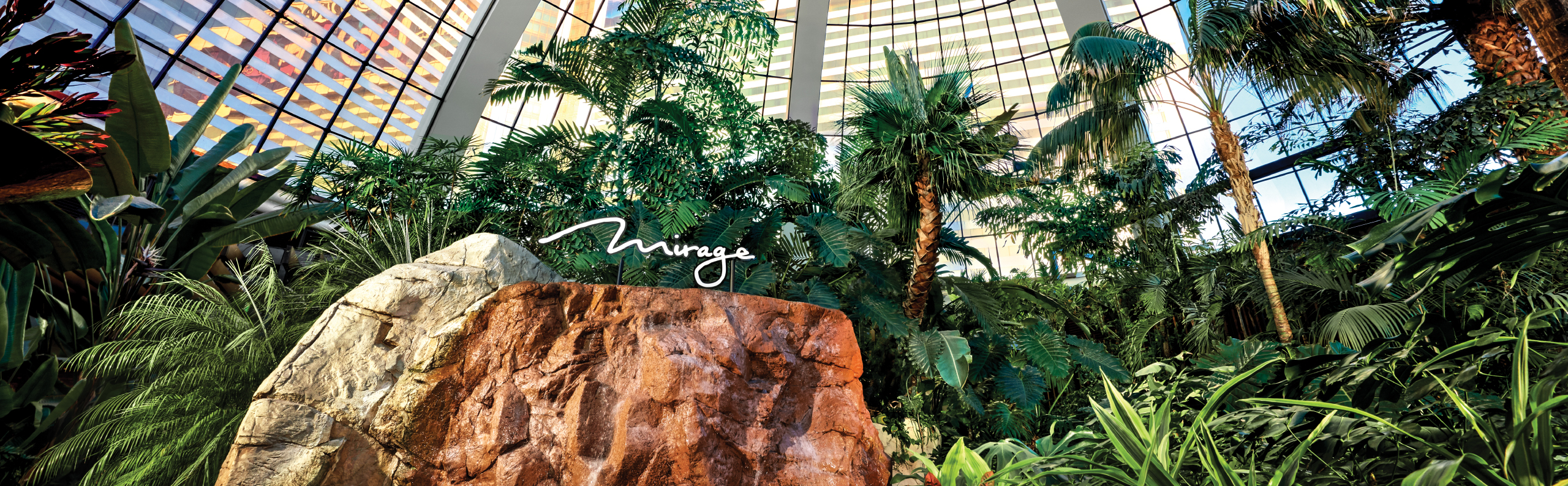 A large rock with the Mirage sign stands among lush indoor tropical plants and tall palm trees under a glass dome at the Braintrust National Conference Center.