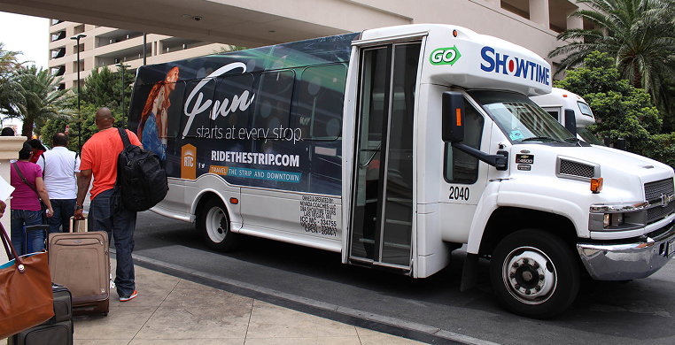 People with luggage wait to board a white Showtime shuttle bus at a hotel entrance with palm trees nearby.