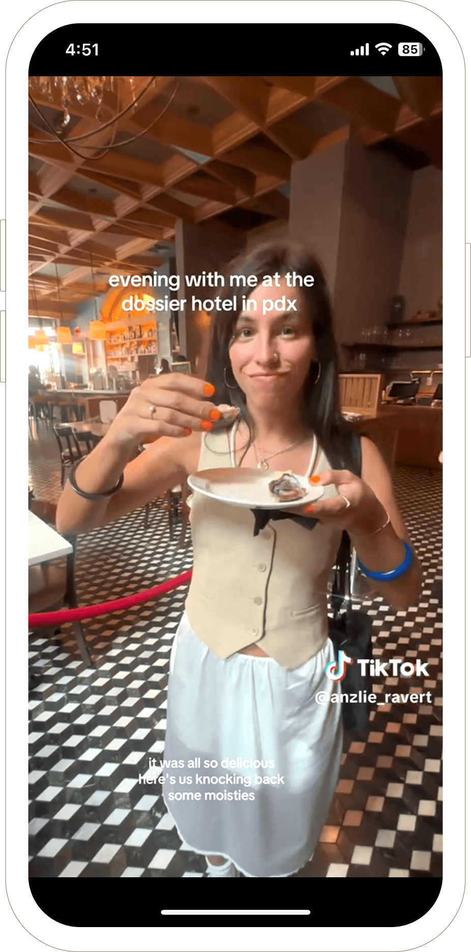 A woman stands in a restaurant holding an oyster and a drink, smiling at the camera.