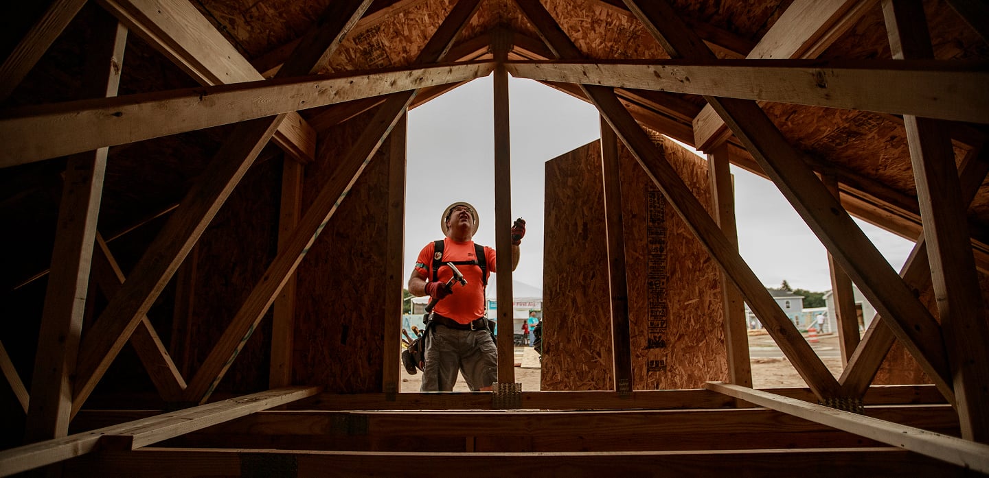A construction worker in an orange shirt stands inside a wooden house frame, looking up at the structure.