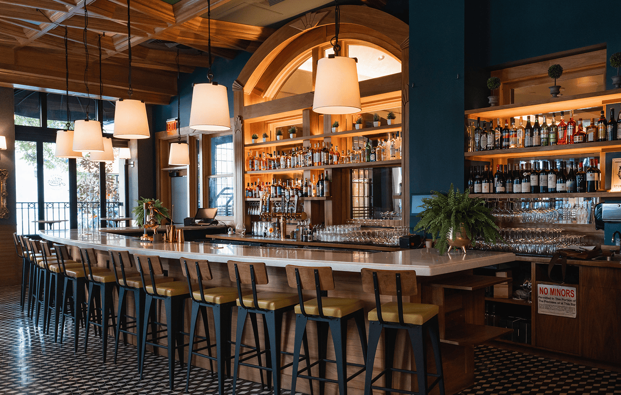 A modern bar with a marble countertop, high chairs, shelves of bottles, and hanging pendant lights.