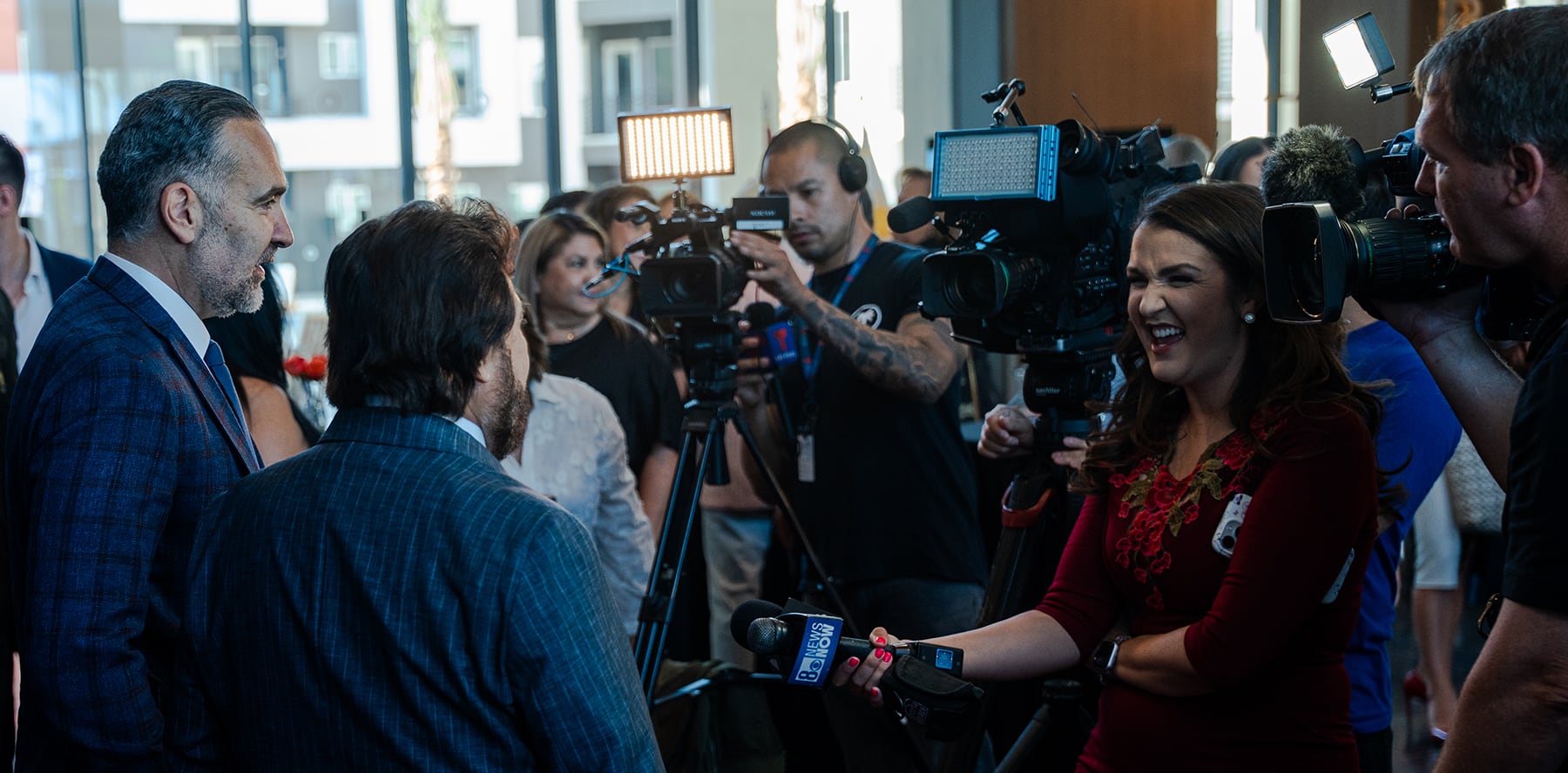 A reporter interviews two men while cameras and media crews capture the event in a busy indoor setting.
