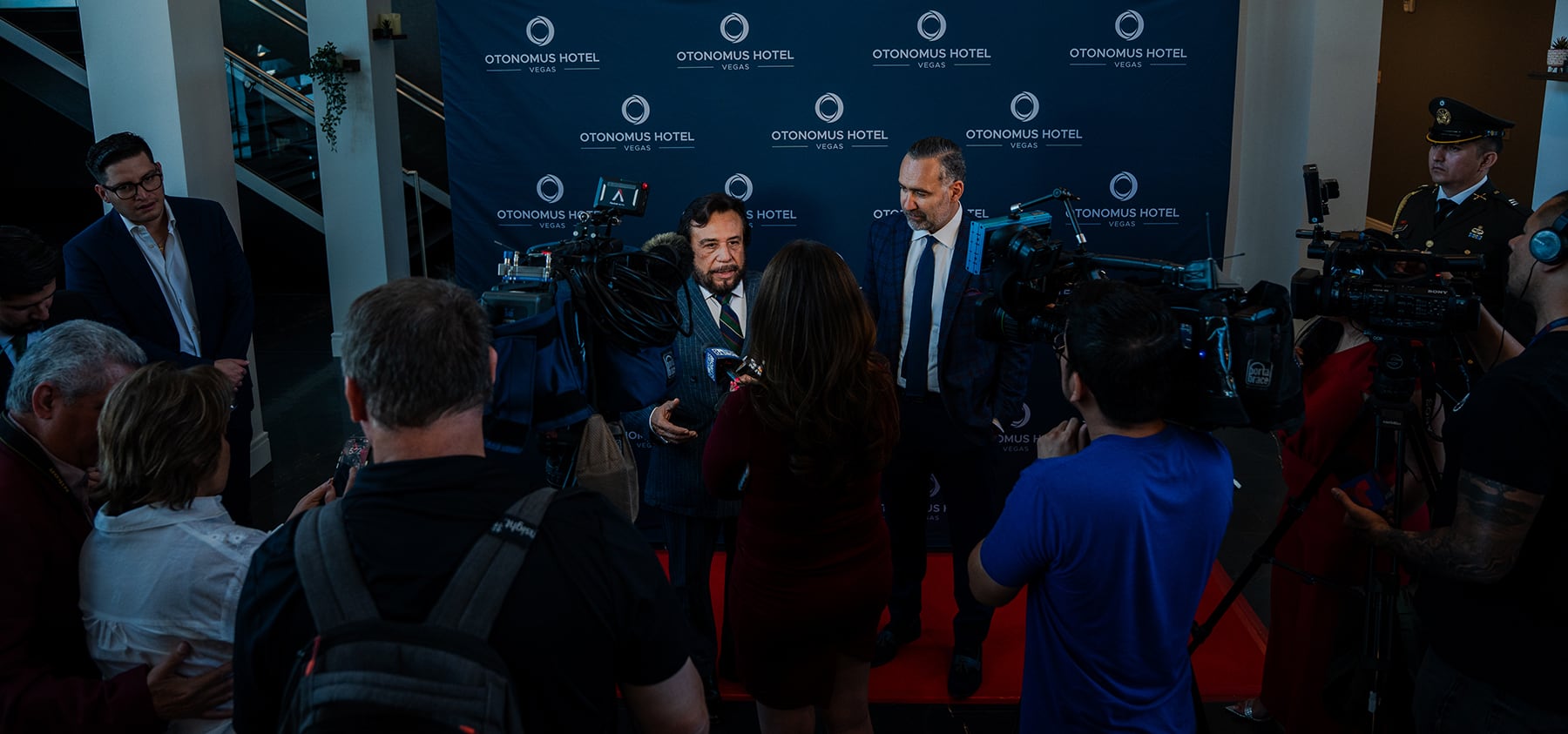 Two men in suits are interviewed by media on a red carpet at an event with a branded backdrop.
