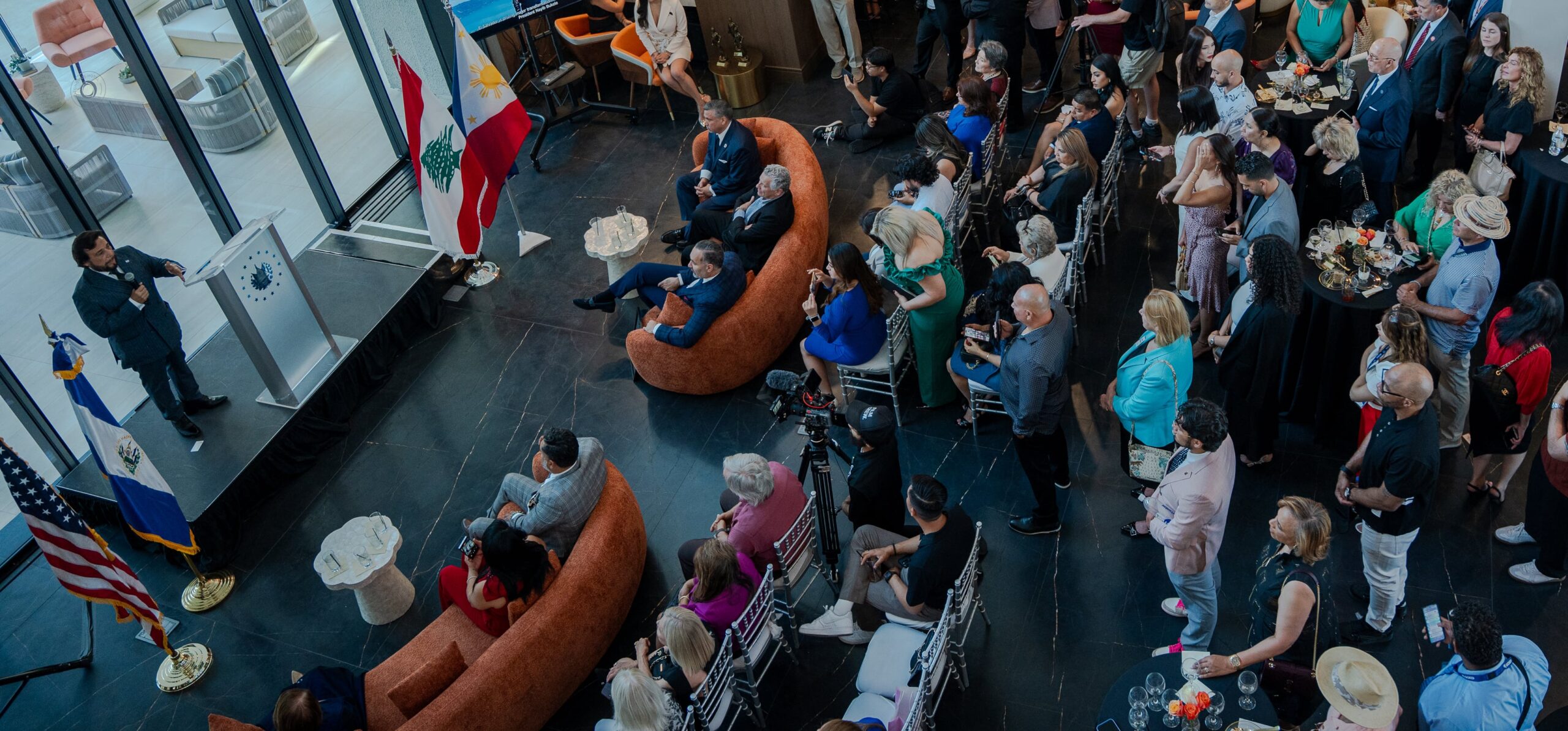 A speaker addresses a seated and standing audience at an indoor event with multiple national flags displayed.