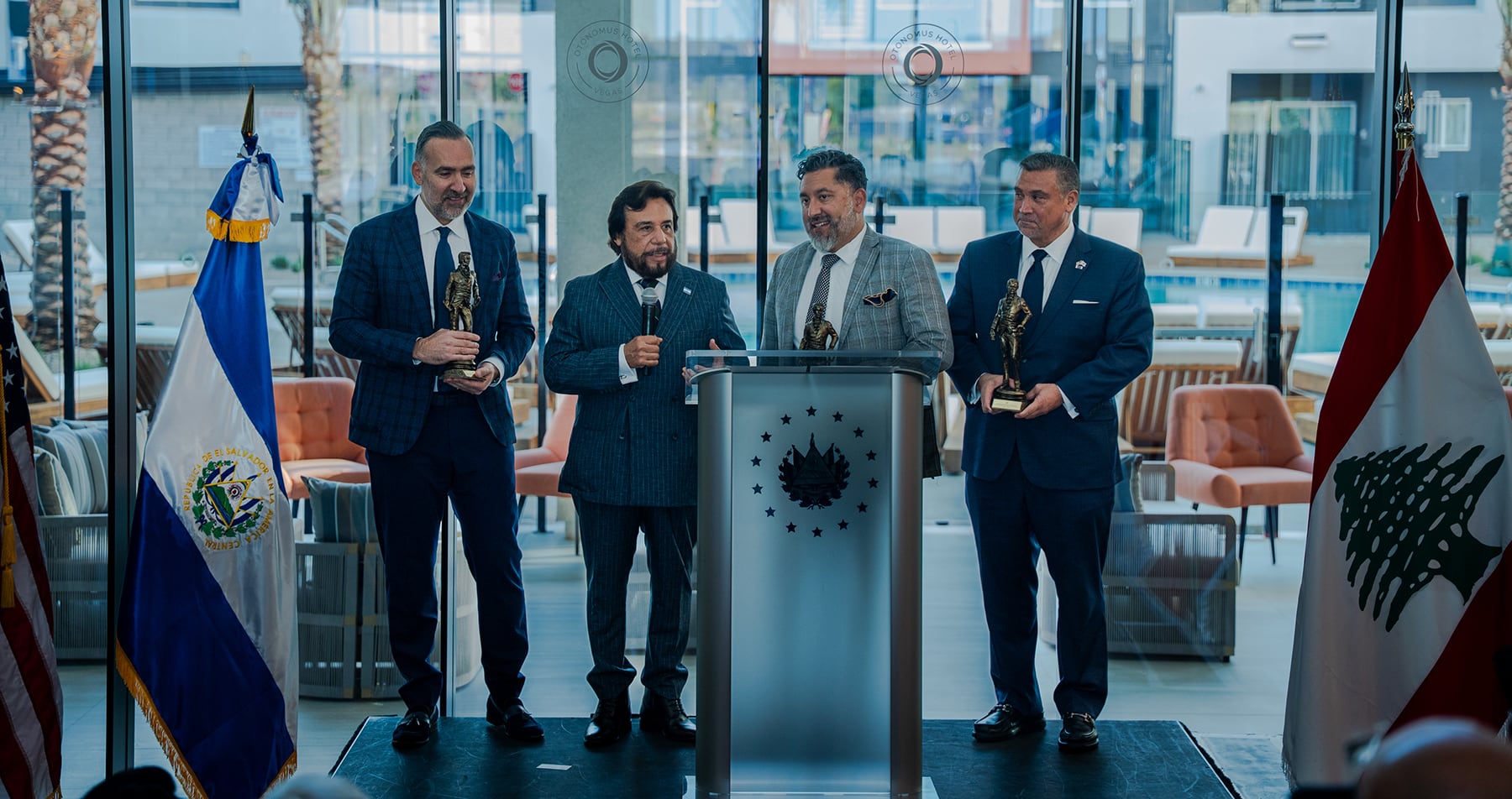 Four men in suits stand with awards at a podium, flanked by El Salvador and Lebanon flags.