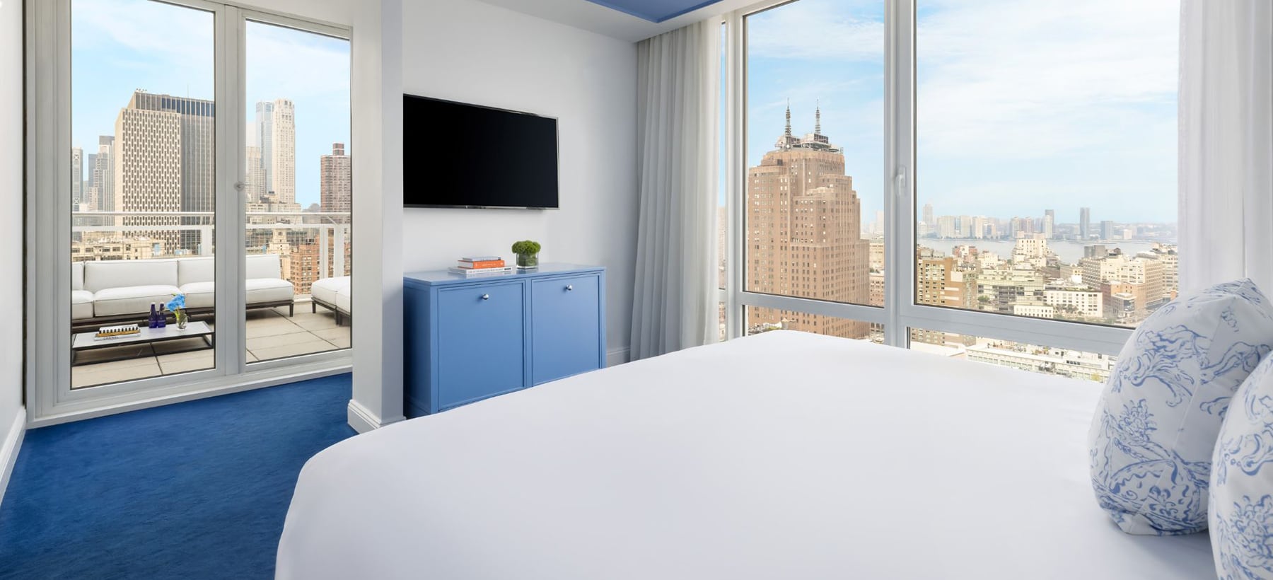 Bright hotel room with city views, large windows, blue carpet, bed, TV, and terrace with outdoor seating.