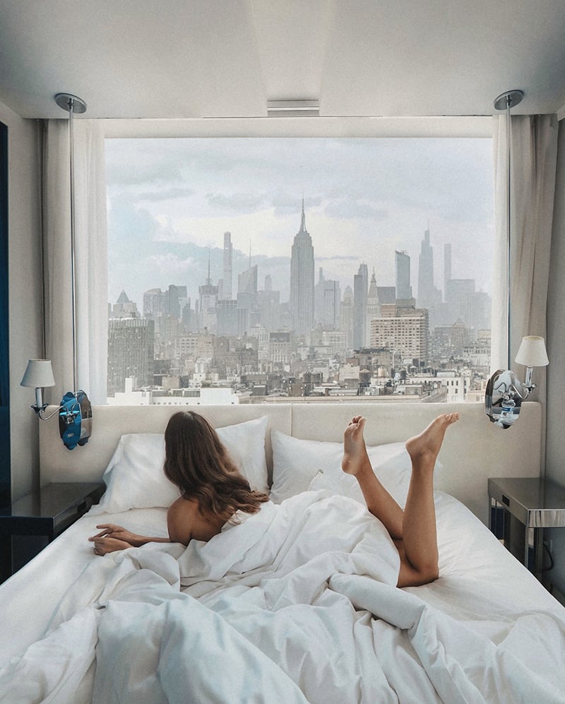 A woman lies on a bed facing a large window with a view of a city skyline on a cloudy day.