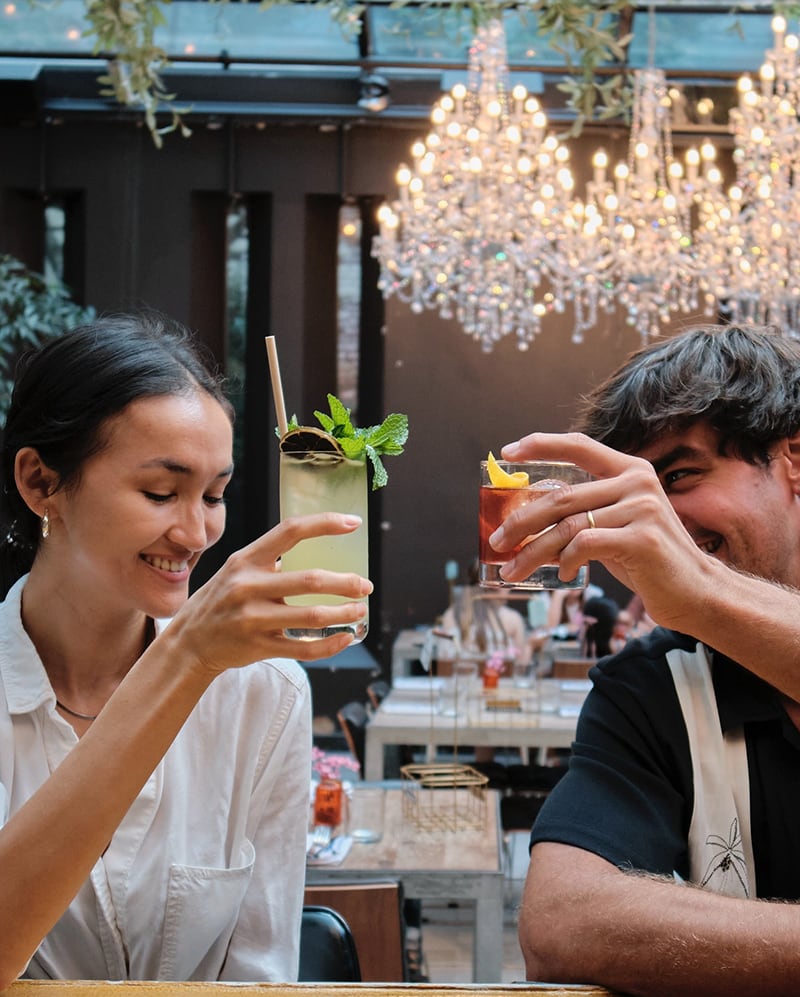 A smiling woman and man toast drinks at a stylish, chandelier-lit restaurant.