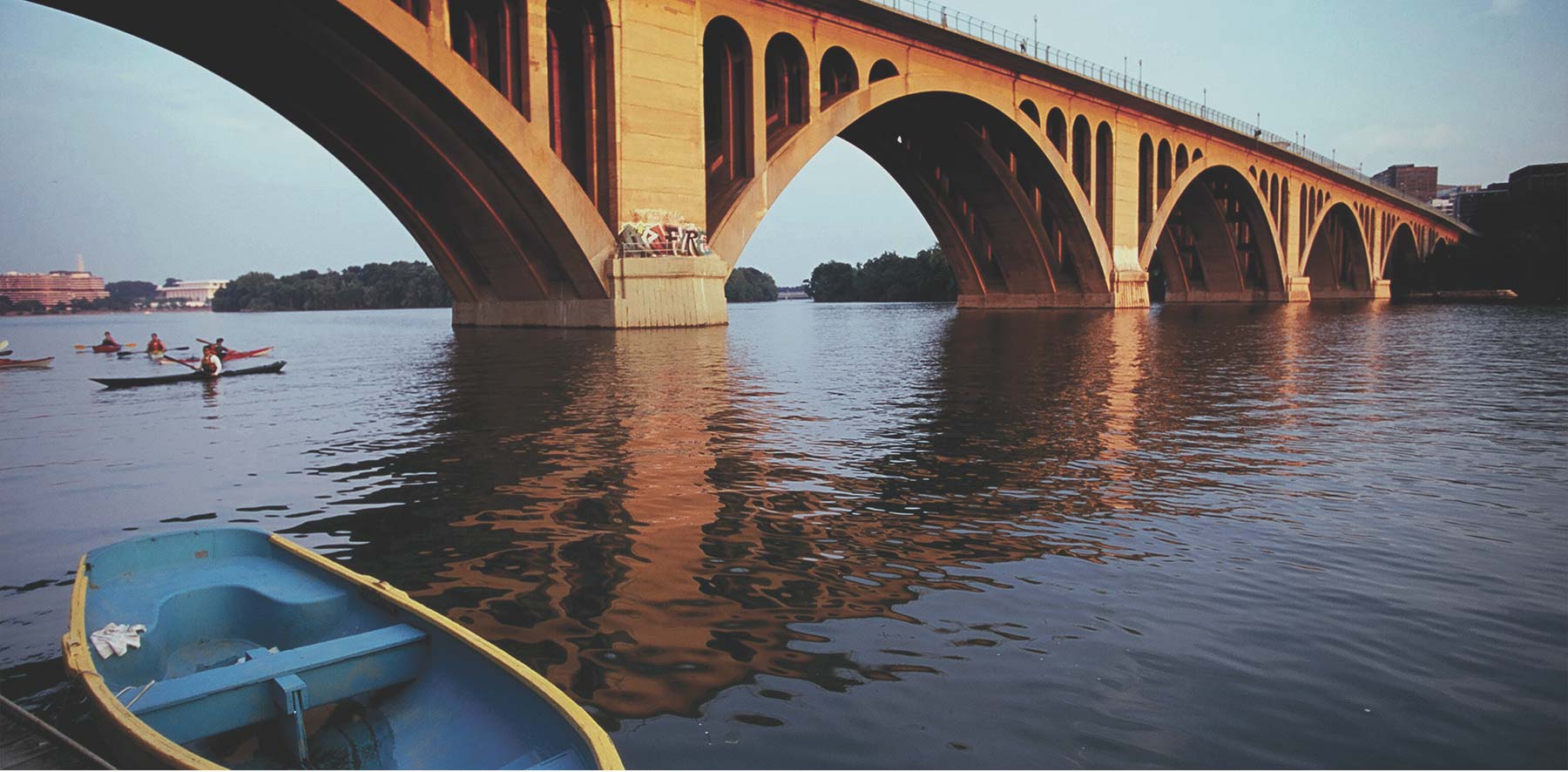 A stone arch bridge over a calm river with kayakers and a blue rowboat in the foreground.