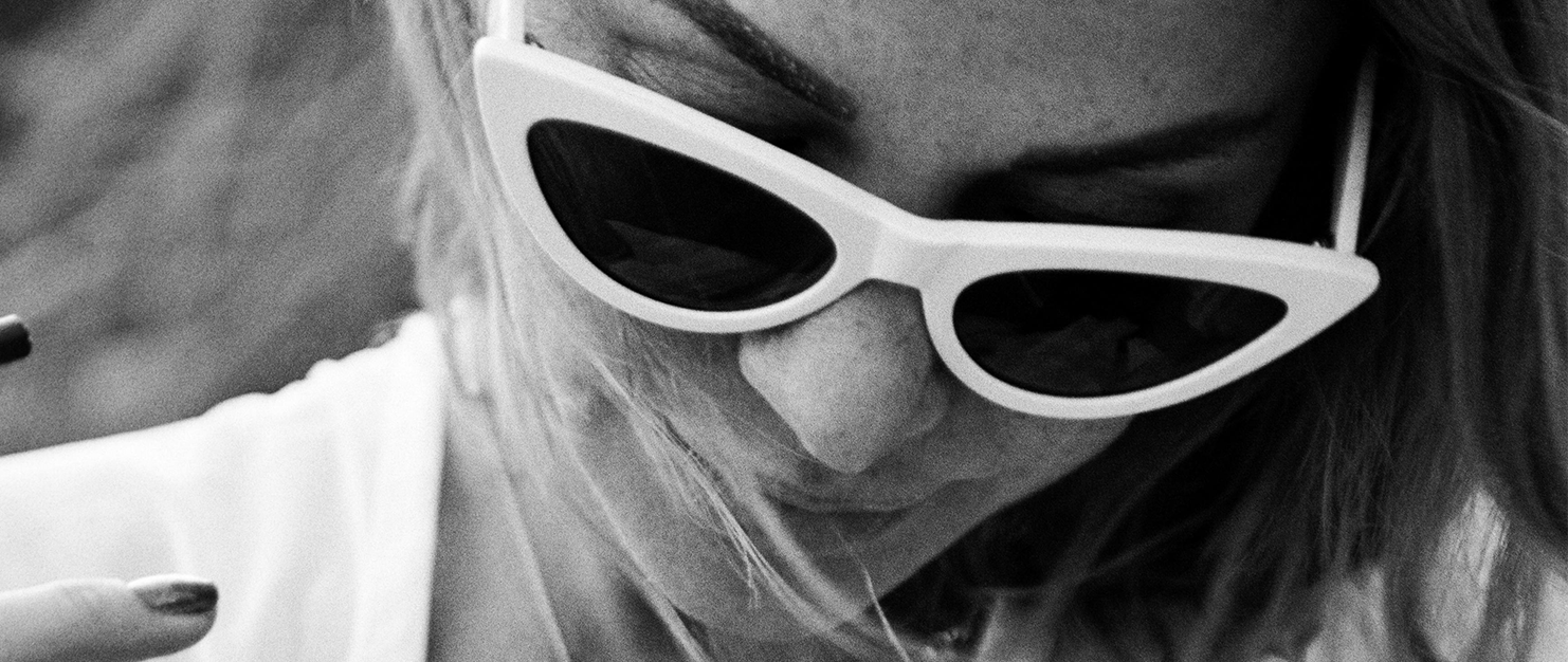 Close-up of a woman wearing cat-eye sunglasses, looking down, with hair partly covering her face. Black and white. A striking visual from our Integrated Marketing & Creative Agency portfolio.
