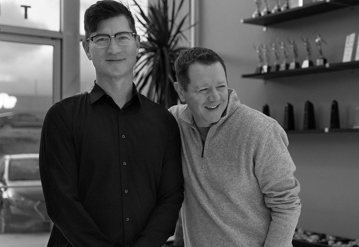 Two men pose together indoors, smiling, with trophies displayed on shelves behind them, reflecting the success and teamwork celebrated at Braintrust Agency - About Us.